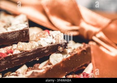 Close-up of artisanal dark chocolate bars topped with dried fruit pieces and nuts, a satin ribbon is visible in the background. Stock Photo