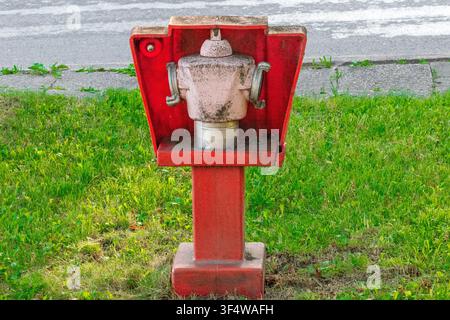 Weathered red metal fire hydrant with open protective cover standing on green grass next to asphalt road and concrete curb dusty white valve head with Stock Photo