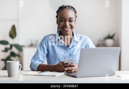 Young Black Female Office Employee Sitting At Workplace With Smartphone In Hands Stock Photo