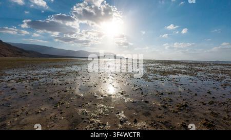 Wide coastal mudflat at low tide with bright sun, scattered clouds, distant mountains and reflections on wet sand, creating a calm open landscape scen Stock Photo