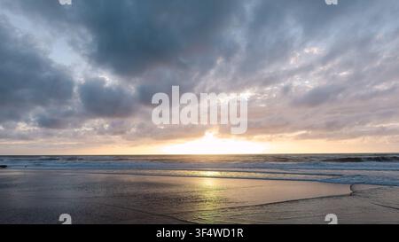 Golden sunset glows on the horizon over an ocean beach, with soft waves rolling onto the wet sand and dark clouds overhead Stock Photo
