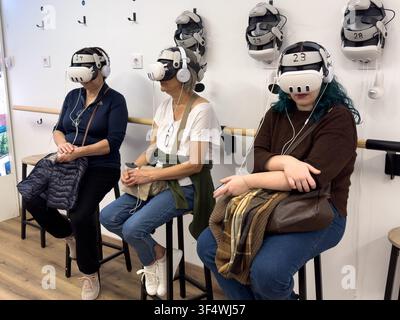 Three seated women wear virtual reality headsets and headphones in a shared immersive technology space. Stock Photo