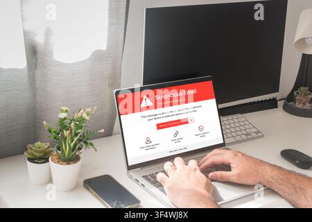 Person types on a laptop showing a suspicious email warning, with a smartphone and desktop monitor on a bright home office desk. Stock Photo