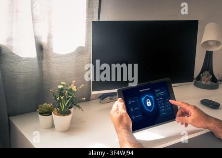 Hands hold a tablet displaying a blue lock and data protection dashboard in front of a desktop monitor in a sunlit home office. Stock Photo