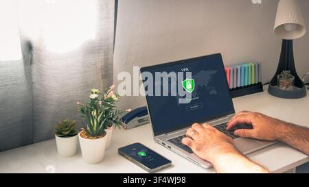 Person works on a laptop while both laptop and smartphone display an active VPN connection with a shield and lock in a home office. Stock Photo