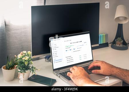 Person types on a laptop displaying a password manager and strong password generator, with a smartphone and desktop monitor in a home office. Stock Photo