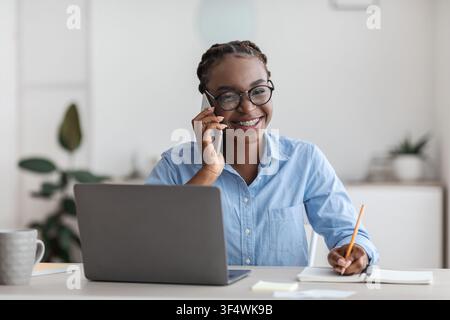 Office Trainee. Black woman talking on cellphone and taking notes at workplace Stock Photo
