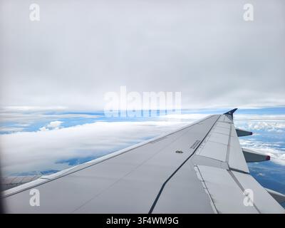 passenger’s window view of an Airbus A320 family aircraft wing soaring gracefully above a thick blanket of clouds under a vast overcast sky. Stock Photo