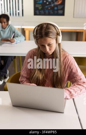 Diverse teen classmates sitting in classroom wearing white headphones and using silver laptop Stock Photo