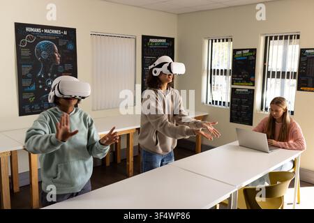 Diverse teenage classmates wearing white VR headsets and interacting at lab tables using laptop Stock Photo