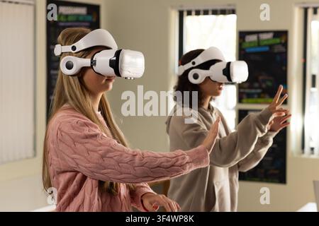 Diverse youth females wearing white VR headsets pink sweater, beige hoodie using VR in tech lab Stock Photo