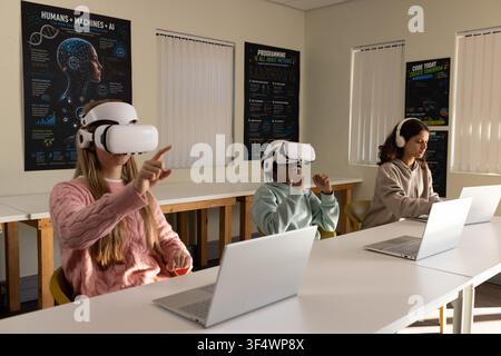 Teenage students wearing pink sweater, beige hoodie using white VR headsets and laptops in tech lab Stock Photo