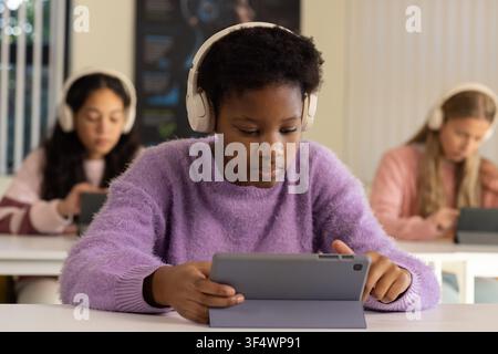 Diverse teen girls in class using tablets and wearing white headphones, center in purple sweater Stock Photo