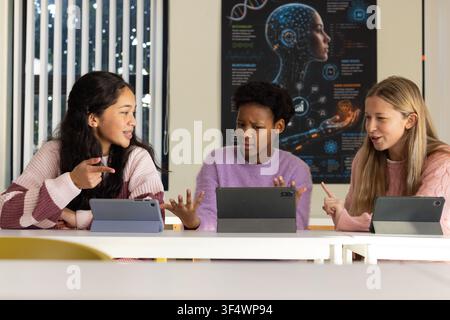 Diverse teenage female students sitting at table in class gesturing at tablets studying AI poster Stock Photo