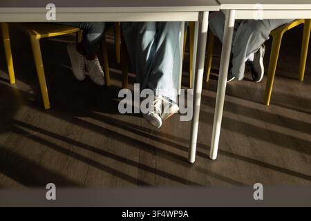 Diverse teenage female students sitting at school tables on yellow chairs showing sneakers, shadows Stock Photo