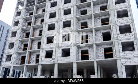 Construction workers are busy on a multi-story building, featuring visible concrete blocks and empty windows. The site is in a dense urban area, refle Stock Photo