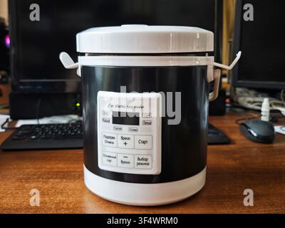 A rice cooker sits on a wooden table with two computer monitors behind it. The cooker has a control panel with buttons for settings and start. Stock Photo