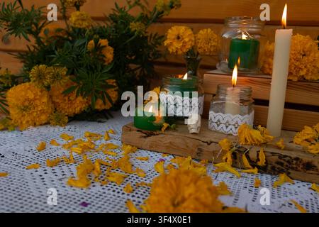 Close view of Day of Dead altar featuring yellow marigold flowers scattered across white lace cloth, lit candles in glass jars, small decorative skull, wooden accents, and warm ritual atmosphere Stock Photo