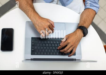 Top view of a man's hands typing on a laptop keyboard at a white round table, with a smartphone nearby. Ideal for concepts of remote work, technology, Stock Photo