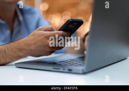 Close-up of a man's hands multitasking with a smartphone and laptop at a desk, featuring a blurred background with warm bokeh lights. Perfect for tech Stock Photo