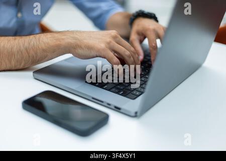 Close-up of a man's hands typing on a laptop keyboard at a white desk, with a smartphone in the foreground. Ideal for remote work, programming, busine Stock Photo