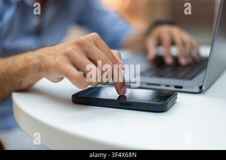 Close-up of a man's hand touching a smartphone screen while typing on a laptop computer. Blurred background, white desk. Great for technology, multita Stock Photo