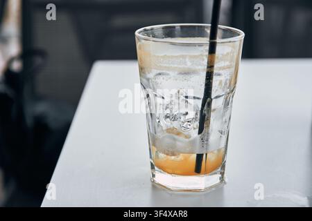 Close-up of a nearly empty tall glass containing melting ice cubes and remaining liquid with a dark drinking straw standing on a bright white table su Stock Photo