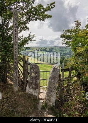 A traditional stone squeeze stile and a public footpath signpost frame a scenic view overlooking the rural village of Bradwell, Peak District Stock Photo