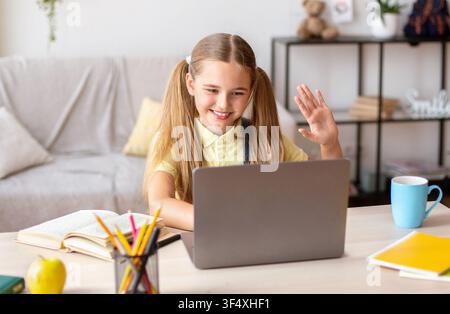 Girl sitting at desk, using laptop and waving at camera Stock Photo