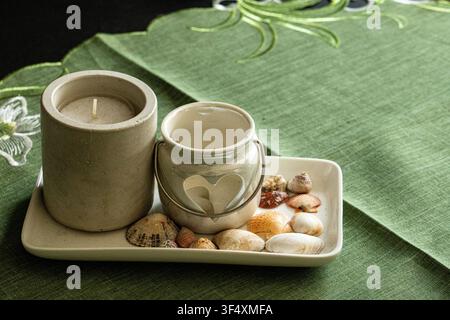 Decorative candles with seashells and pebbles arranged on a tray over a green tablecloth. Relaxing spa and wellness still life. Stock Photo