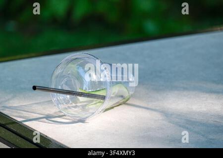Empty disposable plastic takeaway cup with reusable metal drinking straw lying on an outdoor table in natural sunlight. Minimal environmental concept Stock Photo