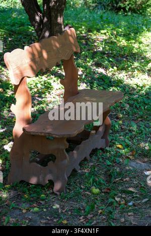 Rustic wooden garden bench standing beneath leafy trees in a peaceful park on a sunny summer day. Traditional outdoor seating surrounded by green gras Stock Photo