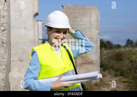 Woman engineer smiling, wearing hard hat and safety vest, holding blueprints at a construction site Stock Photo