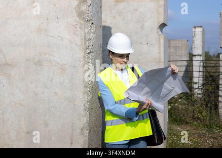 Female architect in safety vest and hard hat reviewing blueprint on site Stock Photo
