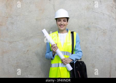 Woman architect engineer wearing a hard hat and safety vest, holding blueprints, looking at camera and smiling Stock Photo
