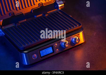 Electric contact grill featuring a digital display, knobs, and textured grilling plates under orange and blue lighting Stock Photo