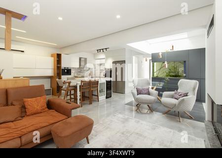 Modern open-plan living room and kitchen interior featuring a leather sofa, plush armchairs, and a large built-in fish tank aquarium under a skylight Stock Photo