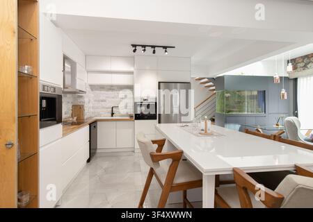 Modern open-plan kitchen and dining area featuring minimalist white cabinetry, stainless steel appliances, a large dining island with wooden chairs Stock Photo