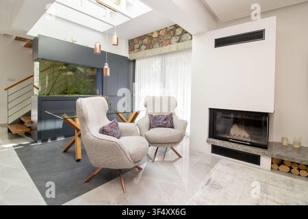 Modern luxury living room interior featuring two plush armchair seats, a contemporary fireplace, a glass dining table, and a custom built-in aquarium Stock Photo
