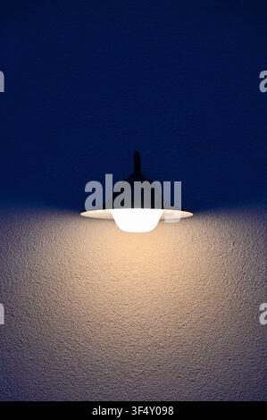 A wall-mounted light fixture illuminates a textured wall in a neutral indoor residential or commercial space, showcasing a frontal view with white and black colors. The metallic fixture. Stock Photo