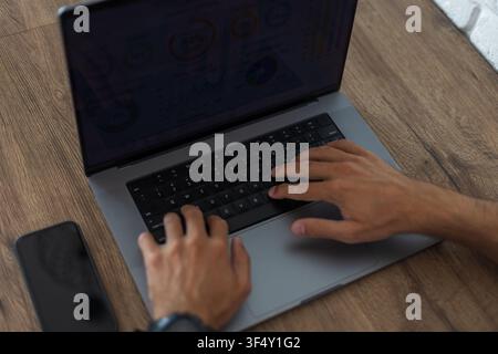 Overhead view of a person's hands typing on a grey laptop keyboard on a dark rustic wooden desk. Modern workspace mockup with a smartphone nearby and Stock Photo