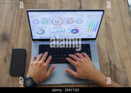 Overhead view of a man's hands typing on a laptop displaying a financial analytics dashboard on a wooden desk. A smartphone mockup lies nearby. Perfec Stock Photo