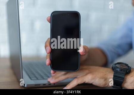 Close-up of a businessman holding a smartphone with a blank black mockup screen in front of an open laptop on a wooden desk. White brick wall blurred Stock Photo