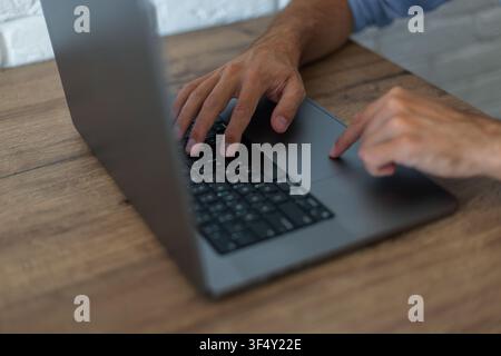 High-angle close-up of a man's hands using a modern laptop on a wooden desk. One hand rests on the black keyboard while a finger touches the trackpad. Stock Photo