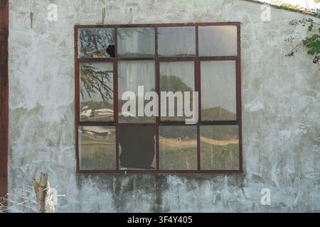 A window with several broken panes sits on the side of a building. The surface shows reflections of trees and the landscape nearby. The scene takes pl Stock Photo