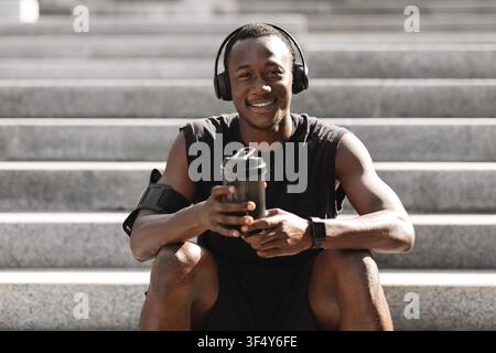 Black Guy Resting After Jogging Outdoors, Listening Music And Having Protein Drink Stock Photo