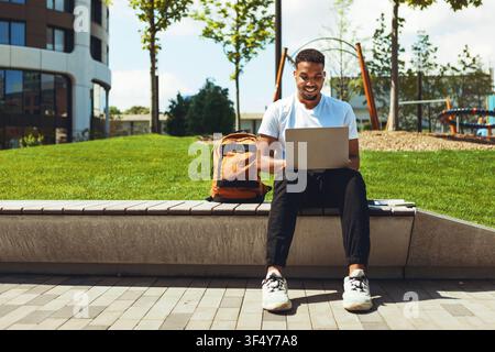 Smiling Young Black Man Working On Laptop Outside, Enjoying Moment In Park Stock Photo