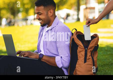 Focused African American Man Working On Laptop In Park With Backpack Nearby, Theft Concept Stock Photo