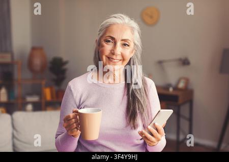 Happy Elderly Caucasian Woman Smiling While Holding Coffee And Smartphone At Home, Wellness Concept Stock Photo