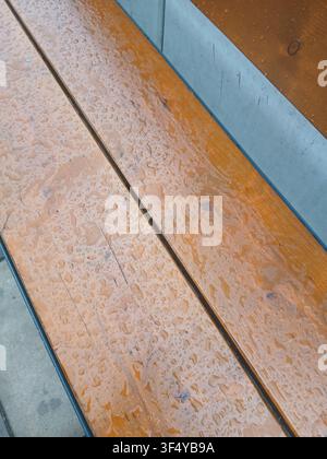 Close up of clear water raindrops on a wet brown wooden bench surface after rain on a city street Stock Photo